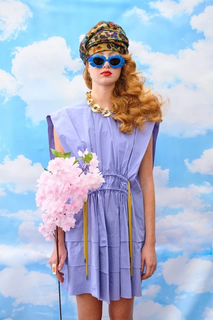 Woman in lilac dress, headscarf, and funky sunglasses holds pink flowers against a cloudy sky backdrop in a fashion editorial.