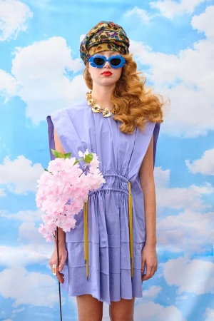 Woman in lilac dress, headscarf, and funky sunglasses holds pink flowers against a cloudy sky backdrop in a fashion editorial.