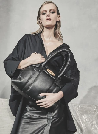 Model with slicked-back hair wearing a black shirt and holding a large black leather handbag against a textured grey background.