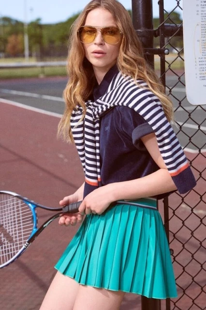 Woman in yellow sunglasses and tennis skirt poses with racket on a tennis court, wearing a navy blue and white striped sweater.