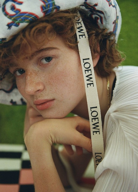 Close-up of a young adult woman with red hair, freckles, and a paisley hat, posing in a white pleated shirt against a green backdrop.