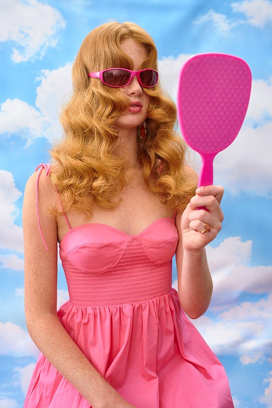 A woman with strawberry blonde hair in a pink dress and sunglasses looks at herself in a pink mirror against a sky backdrop.