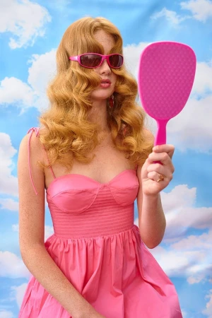 A woman with strawberry blonde hair in a pink dress and sunglasses looks at herself in a pink mirror against a sky backdrop.
