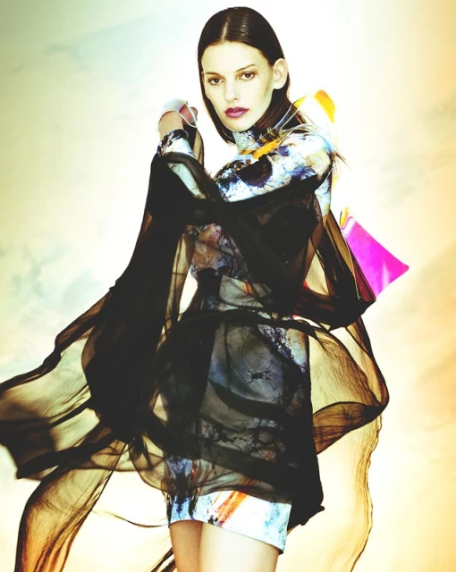 A fashion shot features a slim adult woman in a flowy black dress against a soft, diffused background, blending fashion and art.