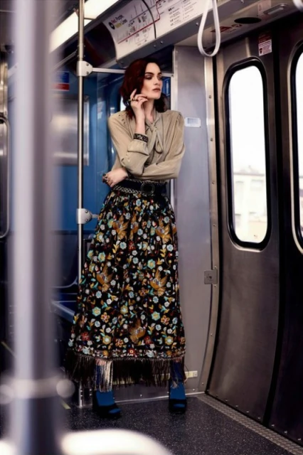 A woman with red hair is wearing a floral print skirt and tan blouse inside a subway car, holding onto the bar on the train.