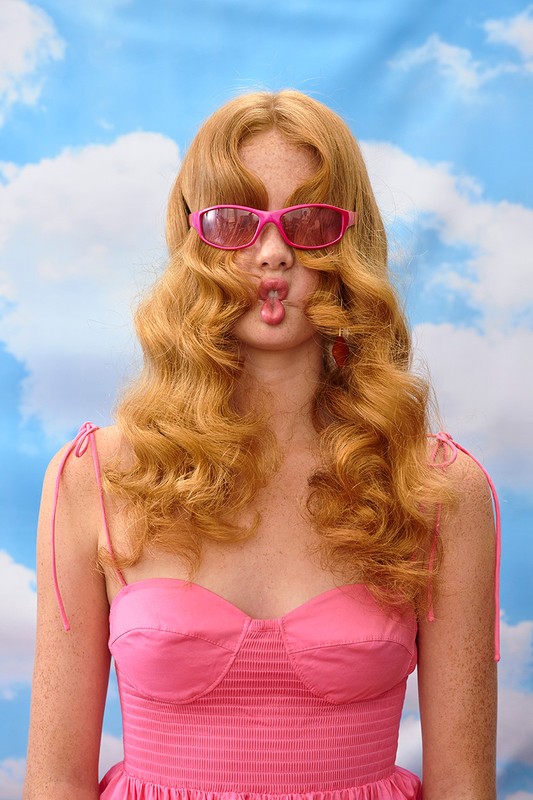 Portrait of a young woman with red hair, wearing pink sunglasses and a pink dress, against a blue sky and white cloud background.