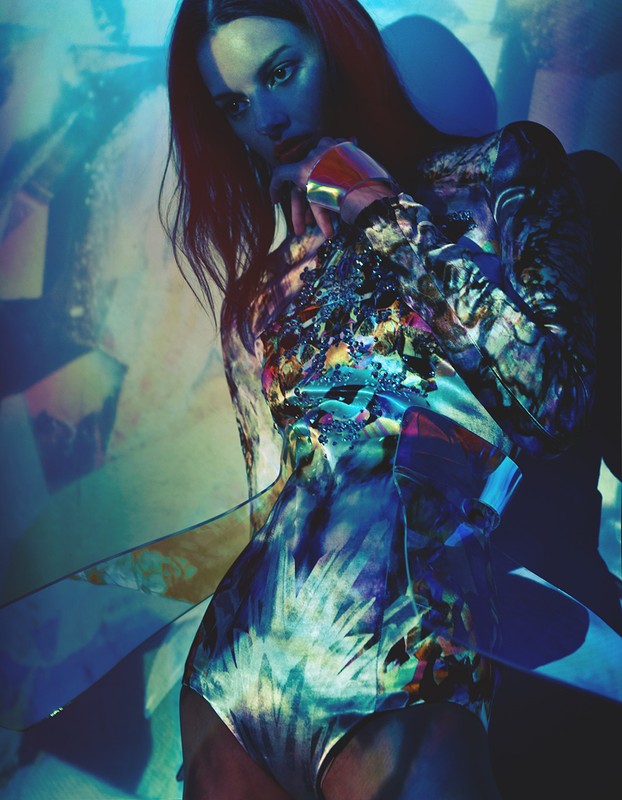 Portrait of a young adult female model in a patterned bodysuit, with a hand to her face and patterned lighting on the wall behind.