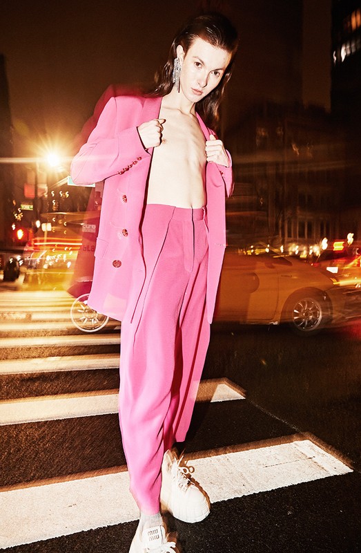 A slender adult poses on a zebra-striped crosswalk at night, wearing a pink pantsuit in an outdoor fashion shot in an urban setting.