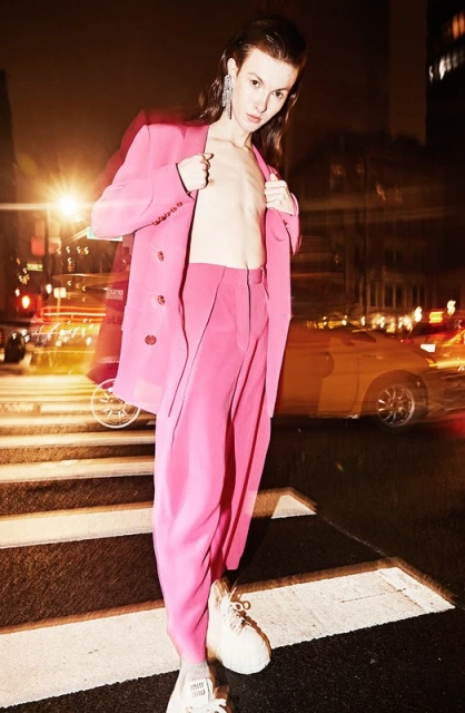 A slender adult poses on a zebra-striped crosswalk at night, wearing a pink pantsuit in an outdoor fashion shot in an urban setting.