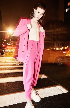 A slender adult poses on a zebra-striped crosswalk at night, wearing a pink pantsuit in an outdoor fashion shot in an urban setting.