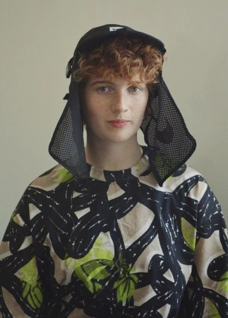 Close-up portrait of a person with curly red hair wearing a black mesh cap and patterned top against a plain wall