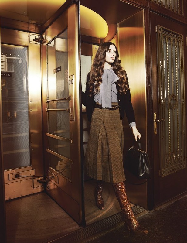 A stylish adult female stands near a bronze door, wearing a grey blouse with a bow, brown skirt, boots, and carrying a black bag.