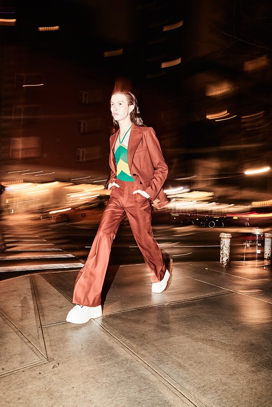 A person in a brown suit walks across a city street at night, showcasing fashion, style, and urban life with blurred background lights.