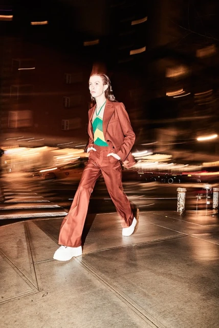 A person in a brown suit walks across a city street at night, showcasing fashion, style, and urban life with blurred background lights.
