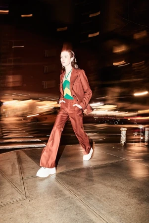 A person in a brown suit walks across a city street at night, showcasing fashion, style, and urban life with blurred background lights.