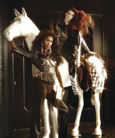 Two female fashion models in shimmering outfits pose next to a white horse statue in a dark photo studio, creating a unique, stylized shot.