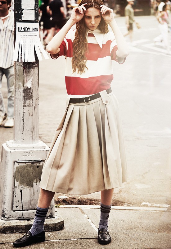 A young adult woman stands on a city street wearing a striped red and white polo shirt and a beige pleated skirt with black loafers.