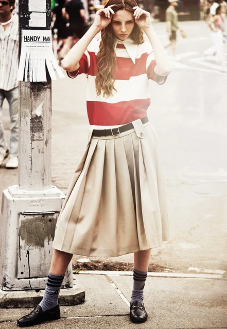 A young adult woman stands on a city street wearing a striped red and white polo shirt and a beige pleated skirt with black loafers.