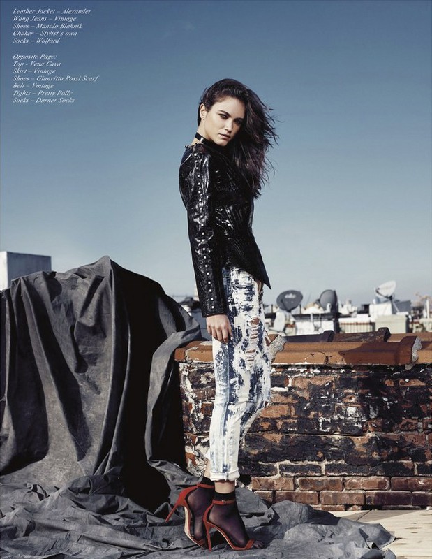 Fashion editorial shot of a young woman in a black leather jacket and acid-wash jeans posing on a rooftop with city buildings in the background.