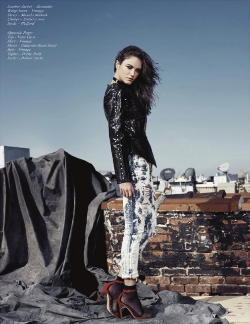 Fashion editorial shot of a young woman in a black leather jacket and acid-wash jeans posing on a rooftop with city buildings in the background.