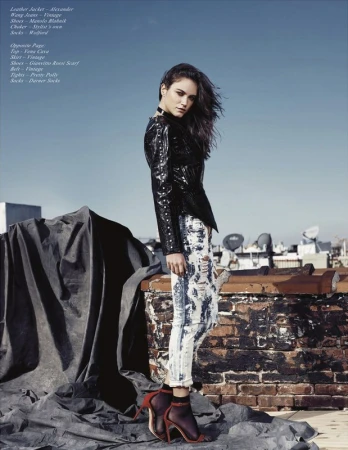Fashion editorial shot of a young woman in a black leather jacket and acid-wash jeans posing on a rooftop with city buildings in the background.