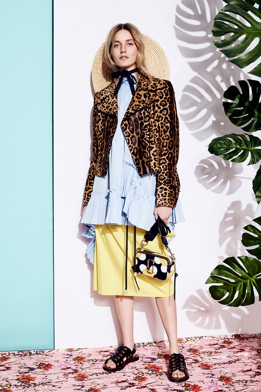 Fashionable woman in animal print jacket and summer hat poses with a handbag against a backdrop of tropical leaves and bright panels.