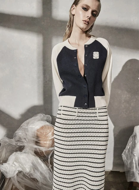 A fair-skinned woman poses indoors wearing a navy and cream cardigan and a striped pencil skirt, in front of plastic-wrapped objects.