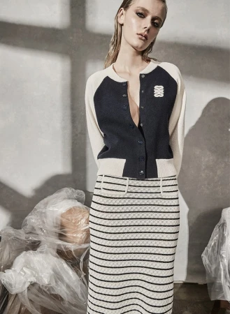 A fair-skinned woman poses indoors wearing a navy and cream cardigan and a striped pencil skirt, in front of plastic-wrapped objects.