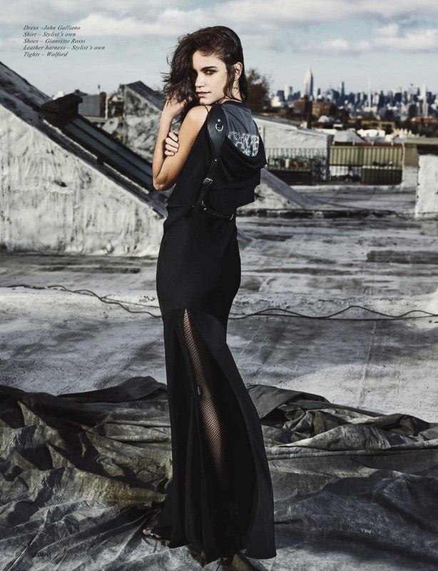 A woman wearing a long black dress and carrying a black backpack poses on a rooftop with a city skyline in the background.