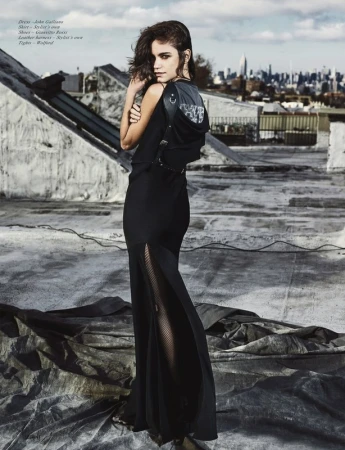 A woman wearing a long black dress and carrying a black backpack poses on a rooftop with a city skyline in the background.
