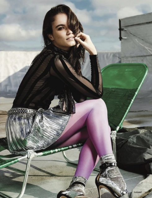 A young woman with dark hair sits on a green chair outdoors, wearing a black top, silver skirt, purple tights, and silver heels.
