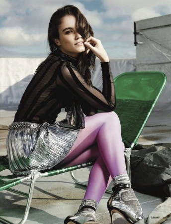 A young woman with dark hair sits on a green chair outdoors, wearing a black top, silver skirt, purple tights, and silver heels.