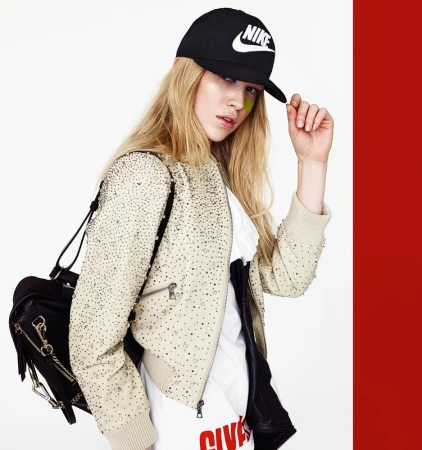 A blonde woman in a studded jacket and Nike cap poses in a fashion shot against a white and red background, showcasing street style.