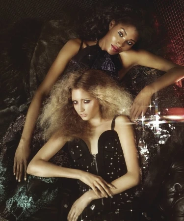 Two women laying down wearing dresses. One woman has blonde curly hair, and the other has dark hair and dark skin, looking at the camera.