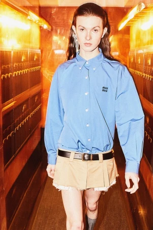 Model wearing a blue button-down shirt and beige mini skirt walking down a hallway with warm lighting, makeup by Fredy Anaya.