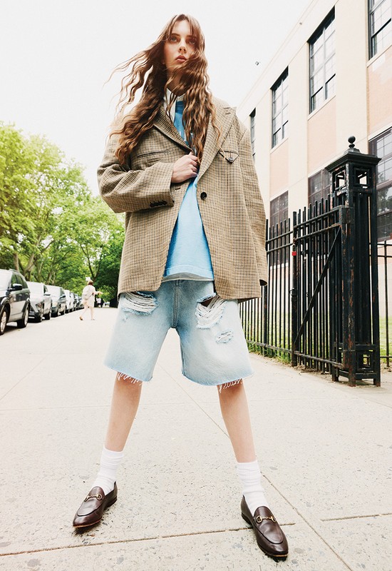 A young woman models on a city street wearing a houndstooth blazer, light blue top, denim shorts, brown loafers, and white socks.