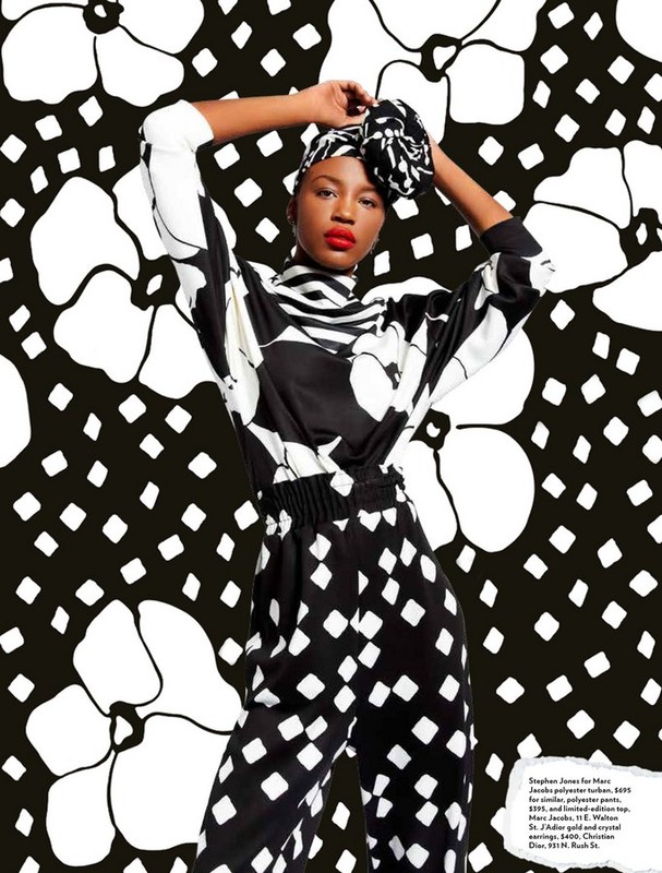 Fashion editorial shot of a Black model wearing a black and white patterned pantsuit and matching head wrap, posing in a studio with a floral backdrop.