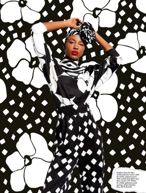 Fashion editorial shot of a Black model wearing a black and white patterned pantsuit and matching head wrap, posing in a studio with a floral backdrop.