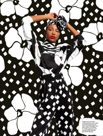 Fashion editorial shot of a Black model wearing a black and white patterned pantsuit and matching head wrap, posing in a studio with a floral backdrop.