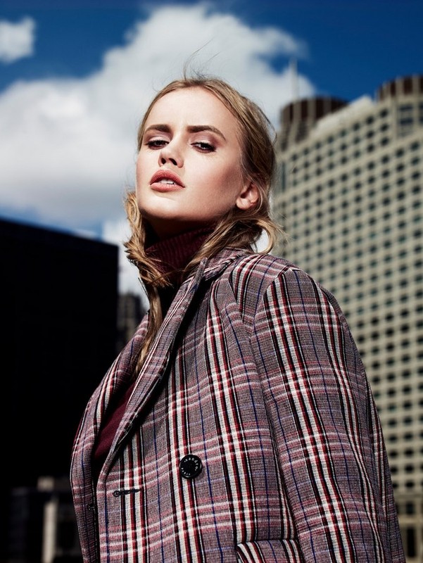 A woman in a plaid coat stands in an outdoor setting against a city skyline, exuding modern fashion and style.