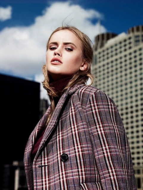 A woman in a plaid coat stands in an outdoor setting against a city skyline, exuding modern fashion and style.