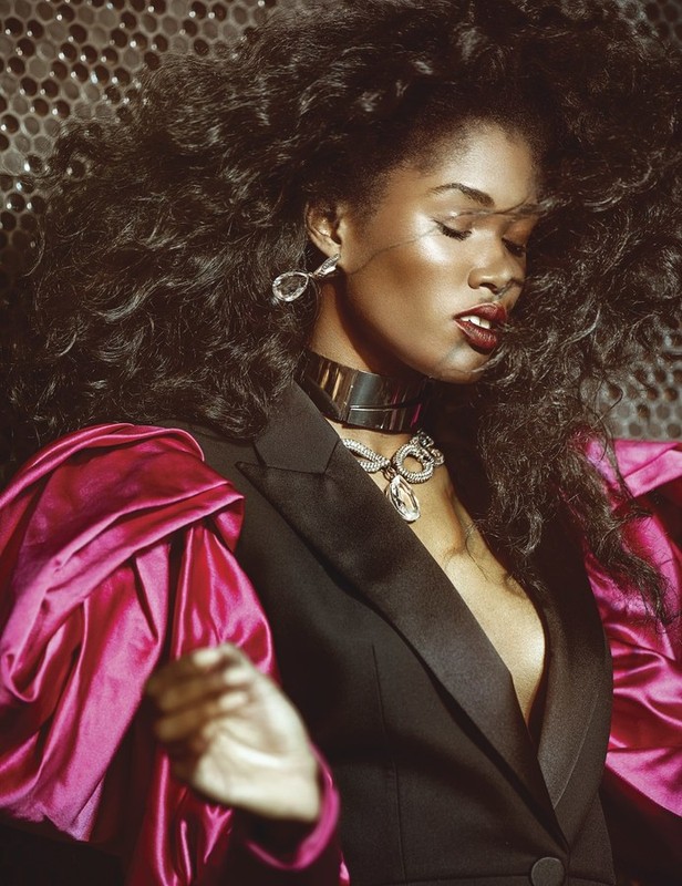 Stunning portrait of a Black woman with voluminous dark hair, wearing statement jewelry, and a black blazer with bright pink sleeves.