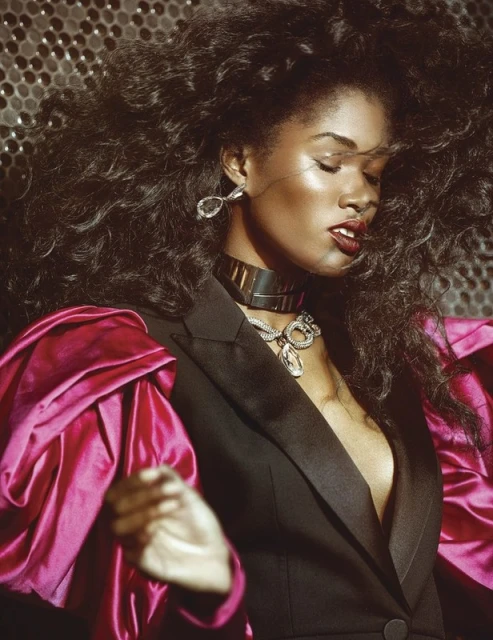Stunning portrait of a Black woman with voluminous dark hair, wearing statement jewelry, and a black blazer with bright pink sleeves.