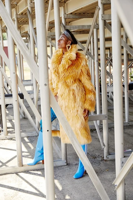A stylish black woman stands in a stadium wearing an orange faux fur coat, blue boots, and hoop earrings, striking a confident pose.