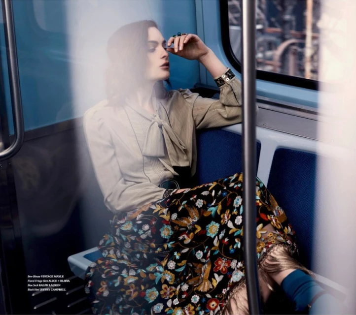 A stylish adult female sits on a subway train in a fashion editorial shot. She wears a floral skirt and a vintage-style blouse.