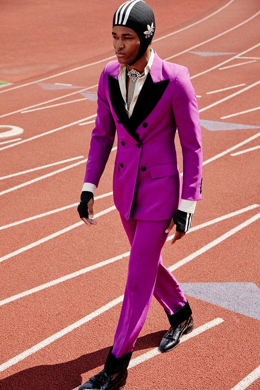 Male fashion model in a purple suit and Adidas beanie walking on a running track with red and white lanes in an outdoor fashion shoot.