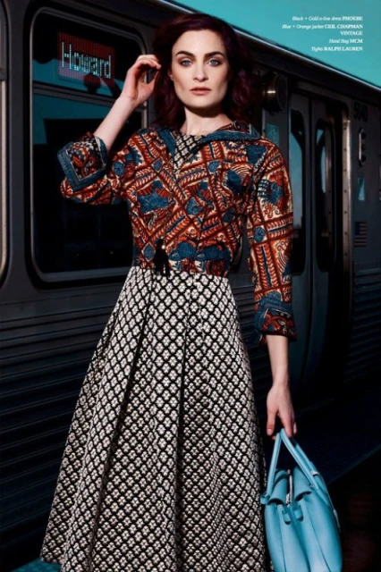 Adult woman poses near a subway car wearing an orange and blue patterned top and black and white patterned skirt holding a blue purse.