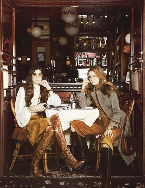 Two women pose at a restaurant table wearing retro 70s fashion, including knee-high boots, suede skirts, and tinted glasses, in a warm, vintage style.