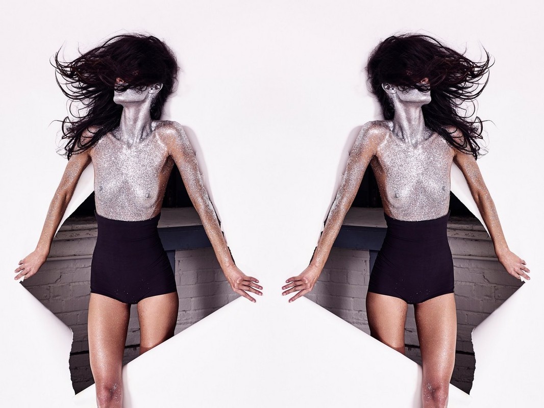 Glamorous mirrored shot of a slender woman covered in silver glitter, partially visible through torn white paper, set against a white backdrop.