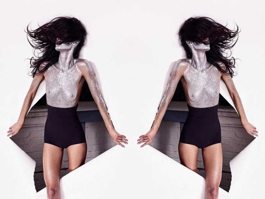 Glamorous mirrored shot of a slender woman covered in silver glitter, partially visible through torn white paper, set against a white backdrop.